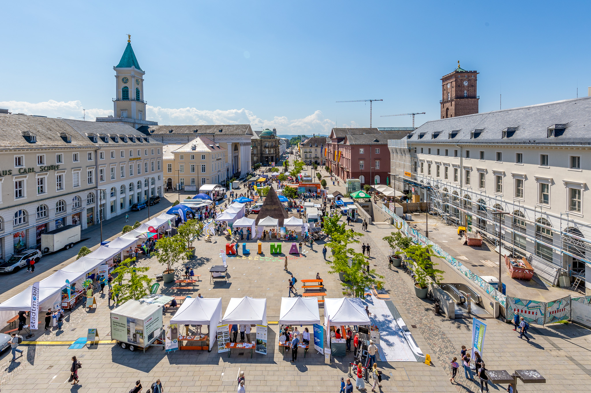 Luftaufnahme des Karlsruher Marktplatzes mit zahlreichen weißen Pavillons, bunten KLIMA-Großbuchstaben und der Stadtpyramide bei blauem Himmel.