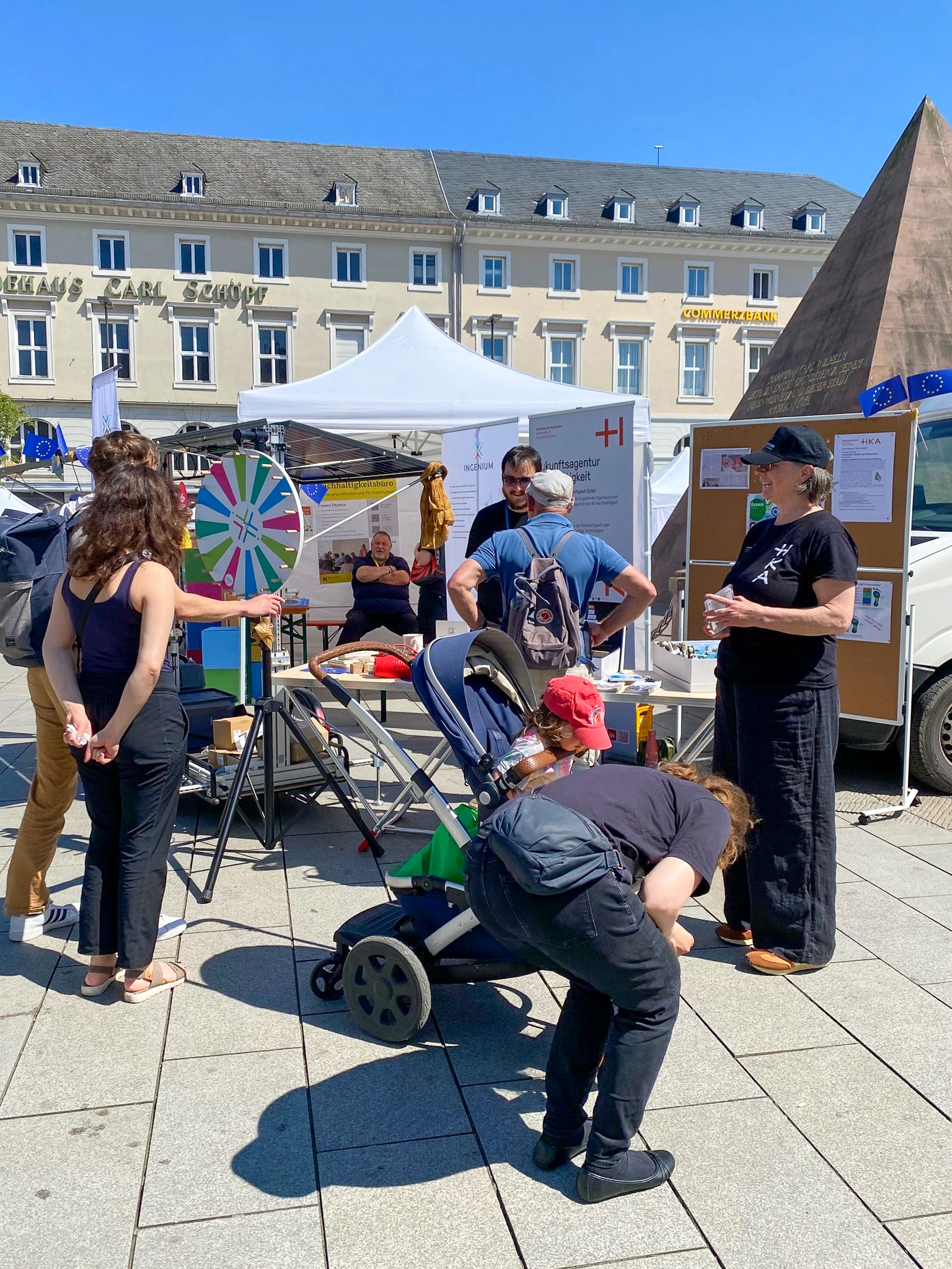Mehrere Personen stehen an einem Ausstellungsstand mit Quizrad und INGENIUM-Rollup unter einem weißen Pavillon auf dem Karlsruher Marktplatz.