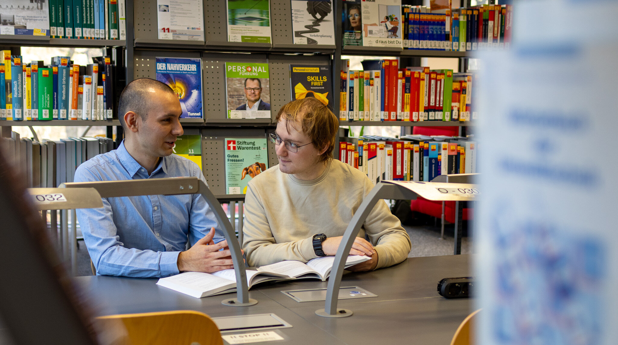 Zwei Studierende sitzen in der Bibliothek und unterhalten sich über ein Buch gebeugt.