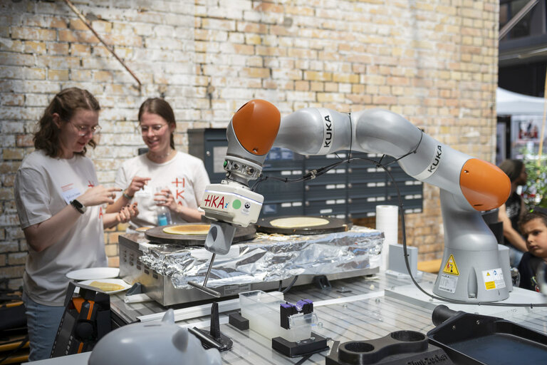 Roboterarm mit der Aufschrift „KUKA“ und „HKA“ bereitet auf einer Crêpe-Platte einen Crêpe zu, im Hintergrund zwei Personen in weißen HKA-T-Shirts vor einer Backsteinwand.