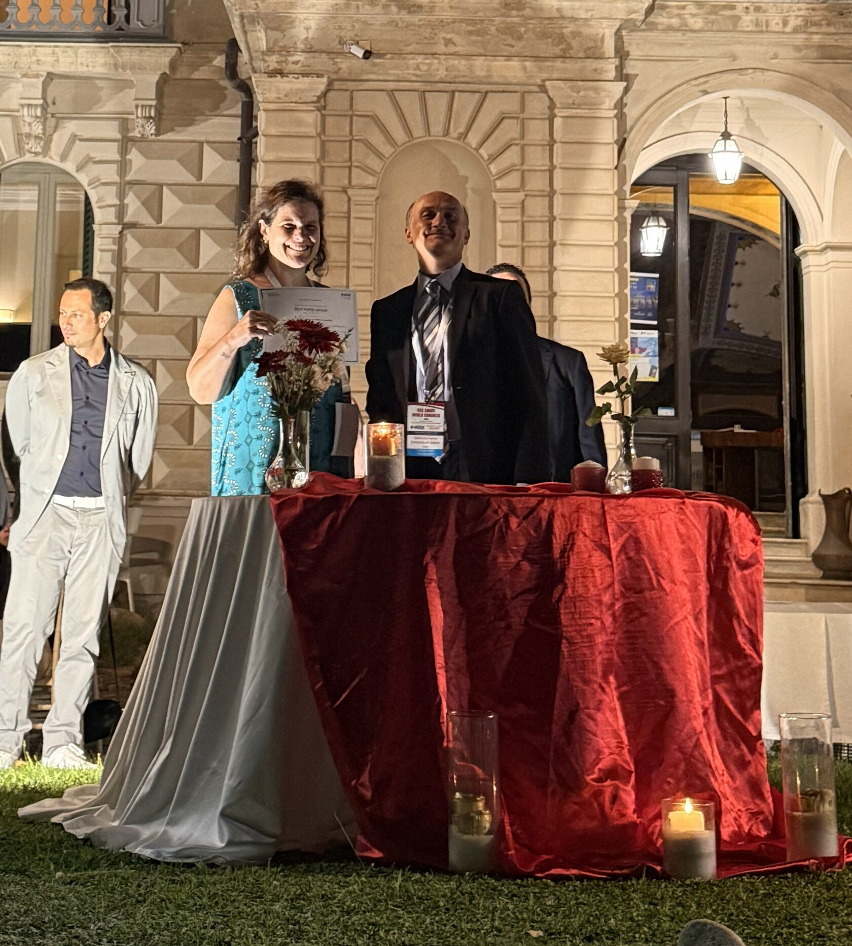 In the photo, Nathalia Sautchuk-Patricio is on the left and Giancarlo Fortino, General Chair of the IEEE Smart World Congress 2026, is on the right. Both are standing in front of a table, and Nathalia Sautchuk-Patricio is holding the award. 