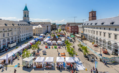 Luftaufnahme des Karlsruher Marktplatzes mit zahlreichen weißen Pavillons, bunten KLIMA-Großbuchstaben und der Stadtpyramide bei blauem Himmel.