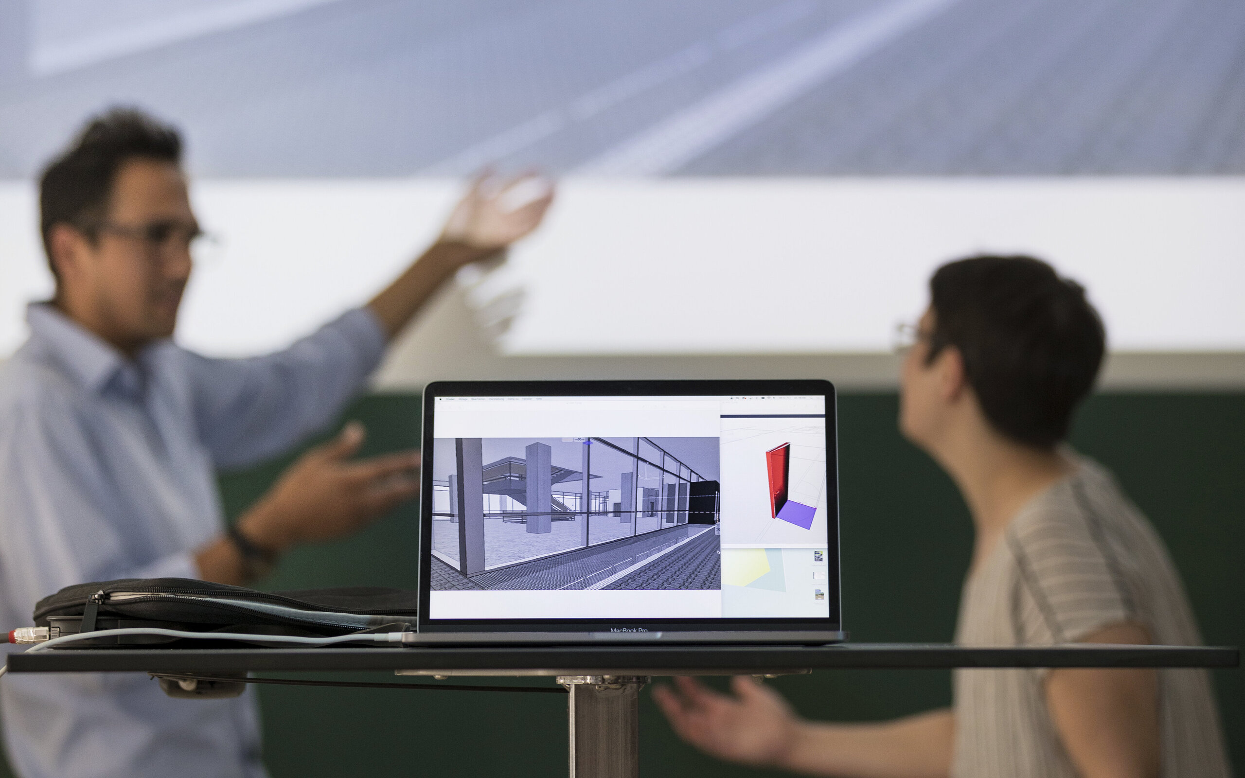 in the foreground a laptop with a building draft, in the background two people discussing in the foreground a laptop with a building draft, in the background two people discussing