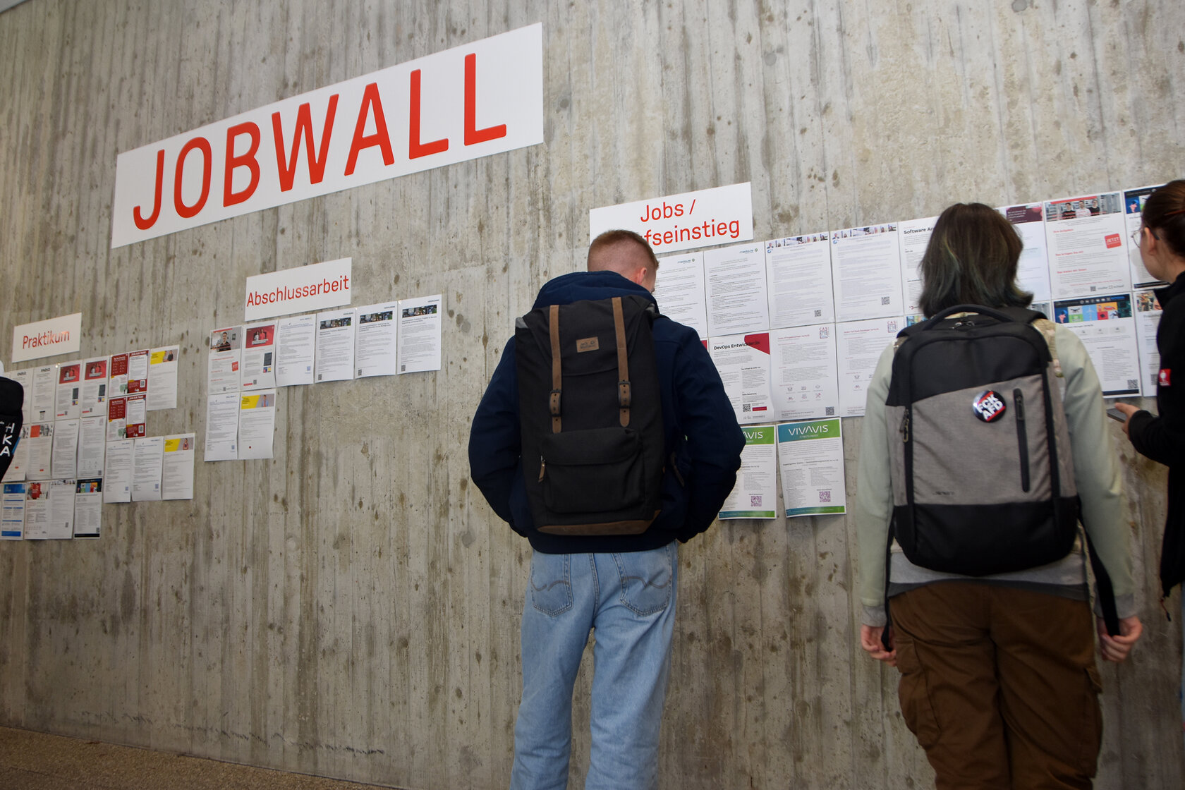 Studierende betrachten zahlreiche Stellenausschreibungen an einer Betonwand. Darüber hängen Schilder mit den Aufschriften „Jobwall“, „Praktikum“, „Abschlussarbeit“ und „Jobs / Berufseinstieg“.