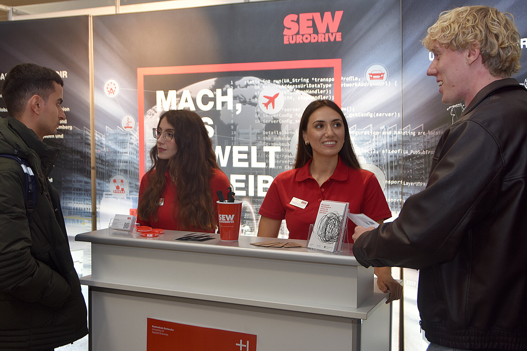 Zwei Studierende unterhalten sich am Messestand von SEW-EURODRIVE mit zwei Mitarbeiterinnen hinter einer Messetheke.