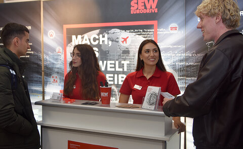 Zwei Studierende unterhalten sich am Messestand von SEW-EURODRIVE mit zwei Mitarbeiterinnen hinter einer Messetheke.
