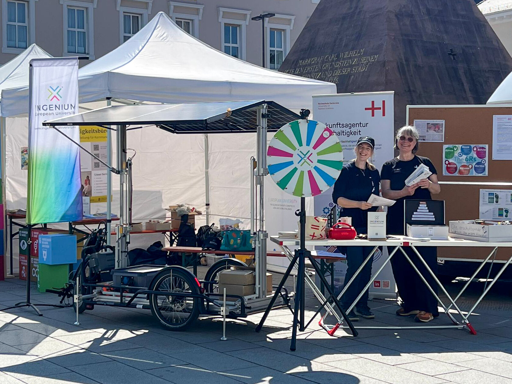 Zwei Frauen in schwarzen HKA-T-Shirts stehen lächelnd hinter einem Ausstellungstisch mit buntem Quizrad, INGENIUM-Banner und einem Lastendreirad im Vordergrund.