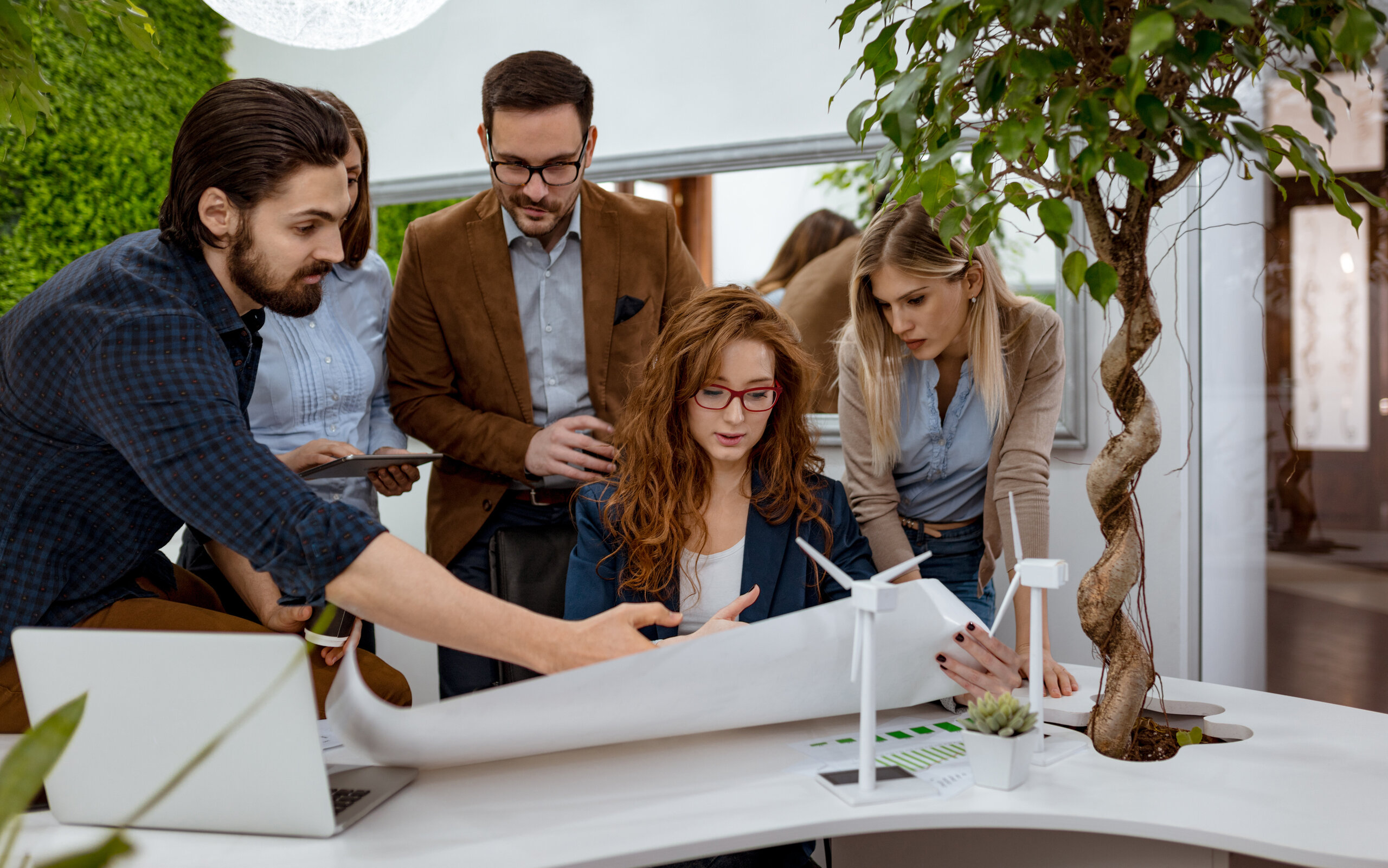 a student working group with laptops and plans is clustered round a table; on this are model wind wheels and a plant a student working group with laptops and plans is clustered round a table; on this are model wind wheels and a plant