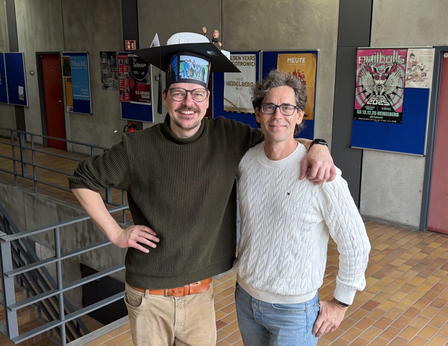 Daniel Hepperle and Matthias Wölfel are standing next to each other with their arms over each other's shoulders. Both are laughing into the camera. Daniel Hepperle is standing on the left and is wearing a cardboard doctor's hat on which, among other things, a 3D model of VR glasses and a 3D model of his VR avatar can be seen; the hat is also covered with pictures of the IIIX team. 