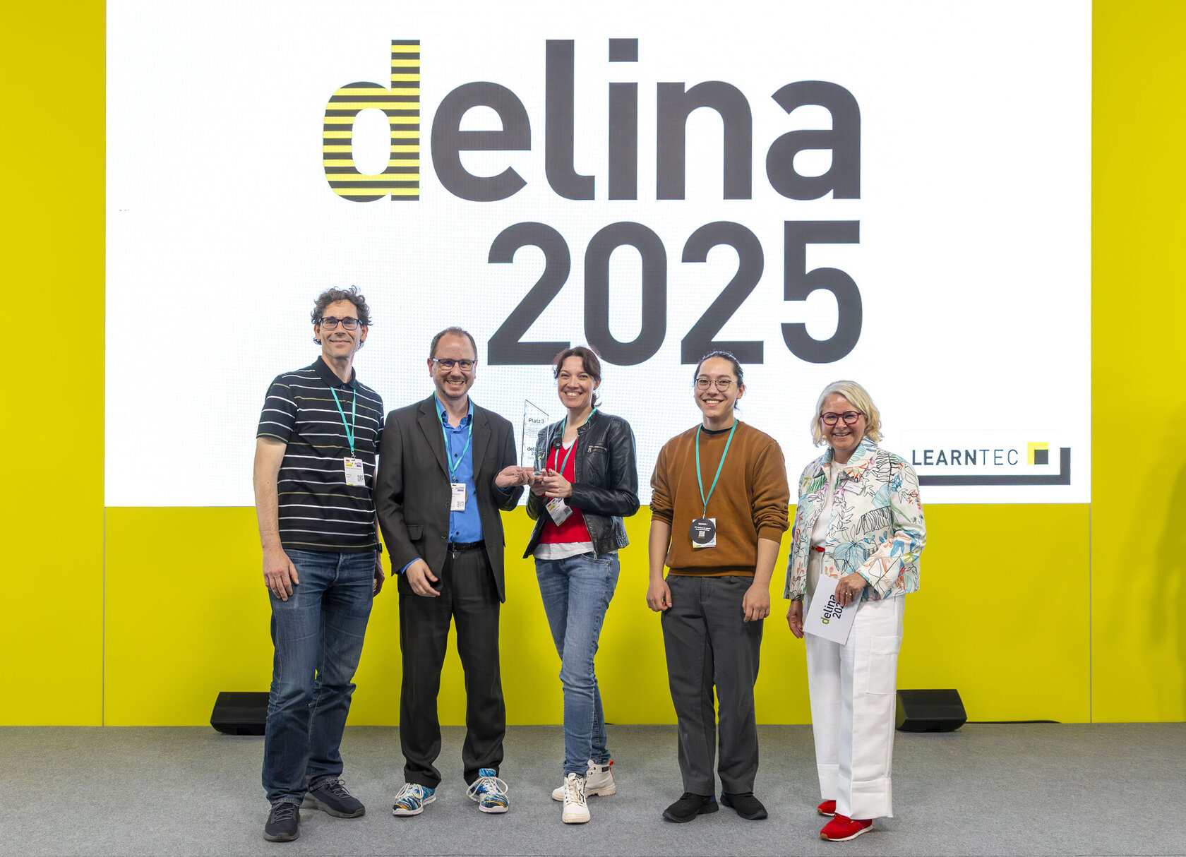 Das Bild zeigt fünf Personen, die auf einer Bühne vor einer großen Leinwand mit der Aufschrift „delina 2025“ stehen. In der Mitte hält Nadine Knobloch die Trophäe. Links von ihr stehen Dr. Oliver Langewitz und Prof. Dr. Matthias Wölfel, rechts Oskar Tanaka und Sünne Eichler. Alle lächeln in die Kamera, der Hintergrund ist hellgelb mit dem LEARNTEC-Logo unten rechts.