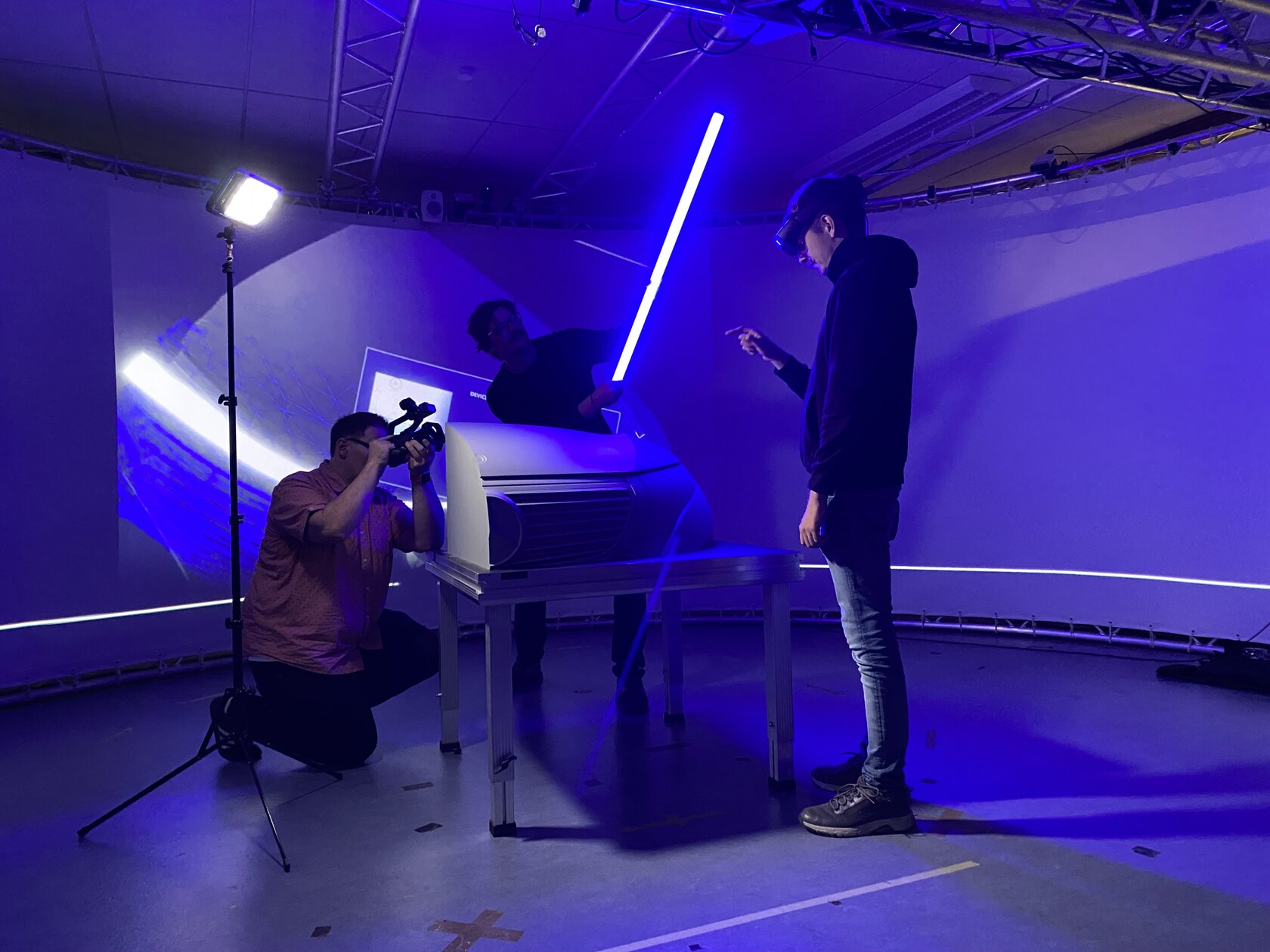 A clay model of a Mercedes Benz can be seen in the center of the picture, surrounded by a cameraman, an employee with the HoloLens and an employee with a halogen light.