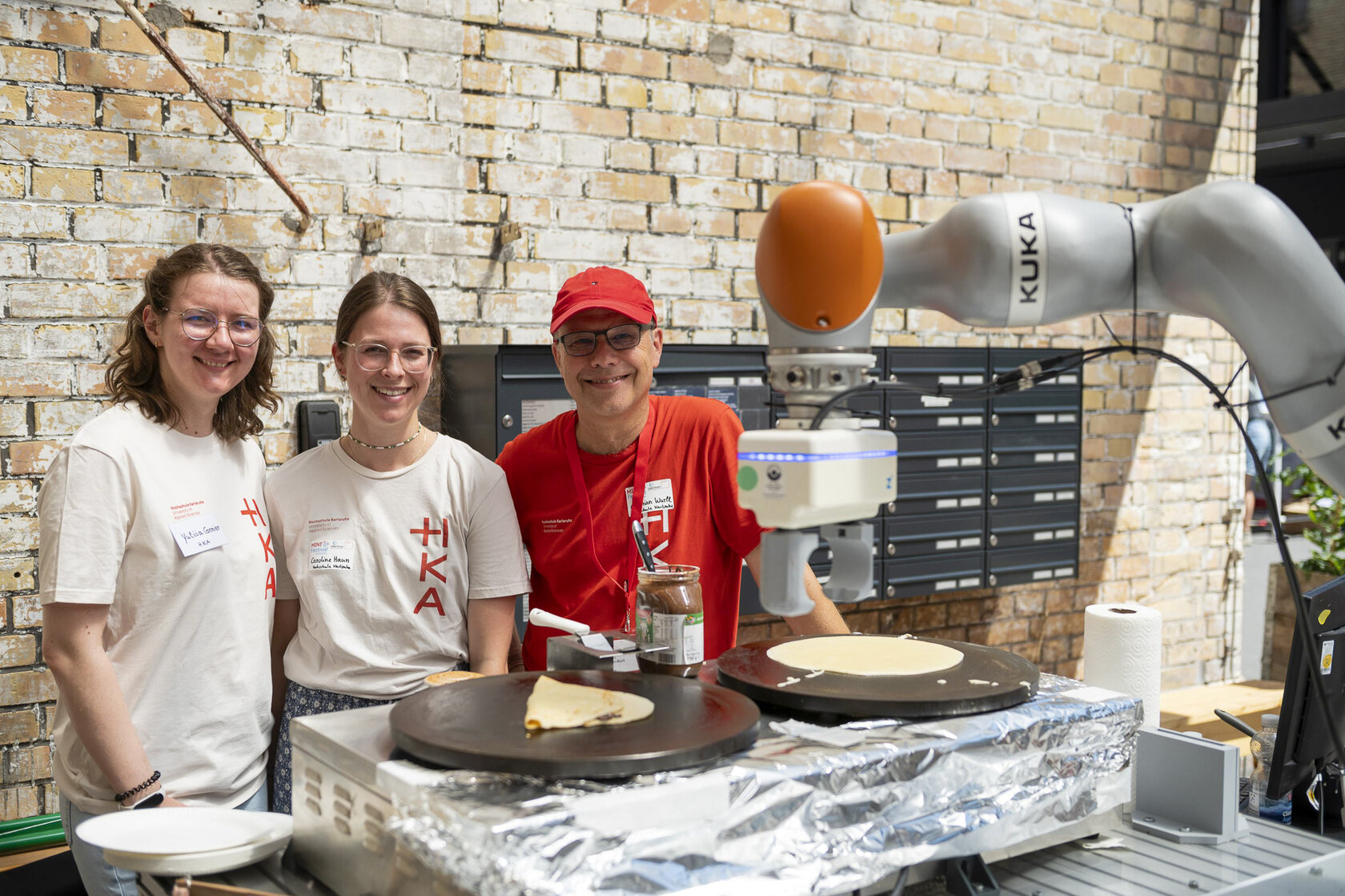 Zwei Frauen und ein Mann in HKA- bzw. MINT-Festival-T-Shirts stehen lächelnd hinter einem Tisch mit Crêpe-Platten und einem Roboterarm vor einer Backsteinwand.