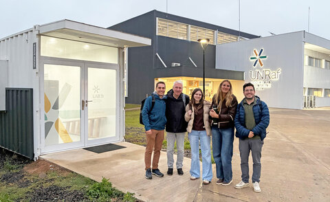 Fünf Personen stehen lächelnd vor einem Laborcontainer mit der Aufschrift "LAB TA" auf dem Campus der UNRaf, im Hintergrund das Hauptgebäude mit UNRaf-Logo.