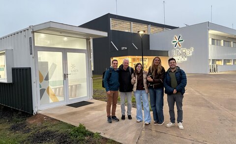 Fünf Personen stehen lächelnd vor einem Laborcontainer mit der Aufschrift "LAB TA" auf dem Campus der UNRaf, im Hintergrund das Hauptgebäude mit UNRaf-Logo.