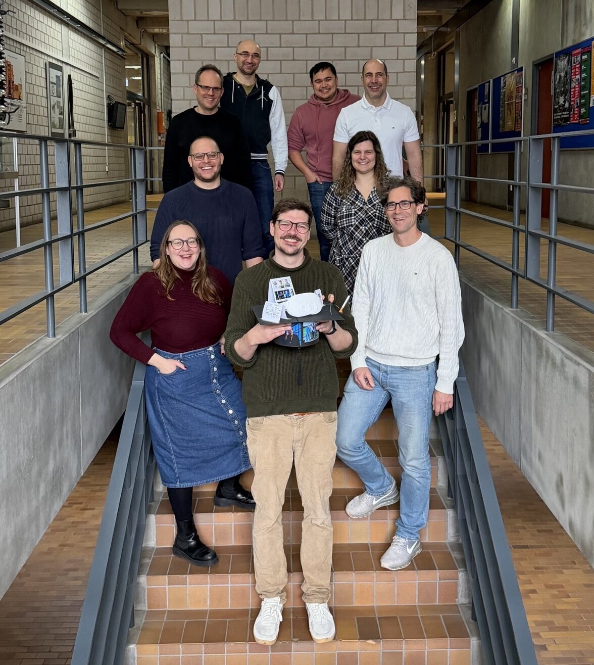 The IIIX team is standing on a staircase, Daniel Hepperle can be seen on the bottom step at the very front. Everyone smiles at the camera. Daniel Hepperle is holding the cardboard doctor's hat made by the team, on which the 3D model of VR goggles and a 3D model of his VR avatar can be seen, among other things; the hat is also covered with pictures of the IIIX team. 