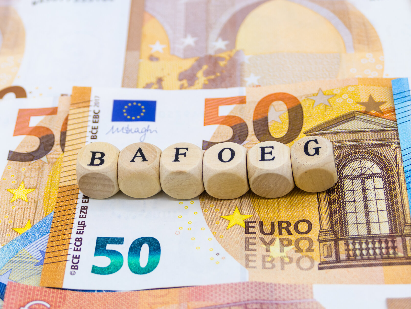 dice with the letters of the word "BAFOEG" on them lying on some banknotes dice with the letters of the word "BAFOEG" on them lying on some banknotes