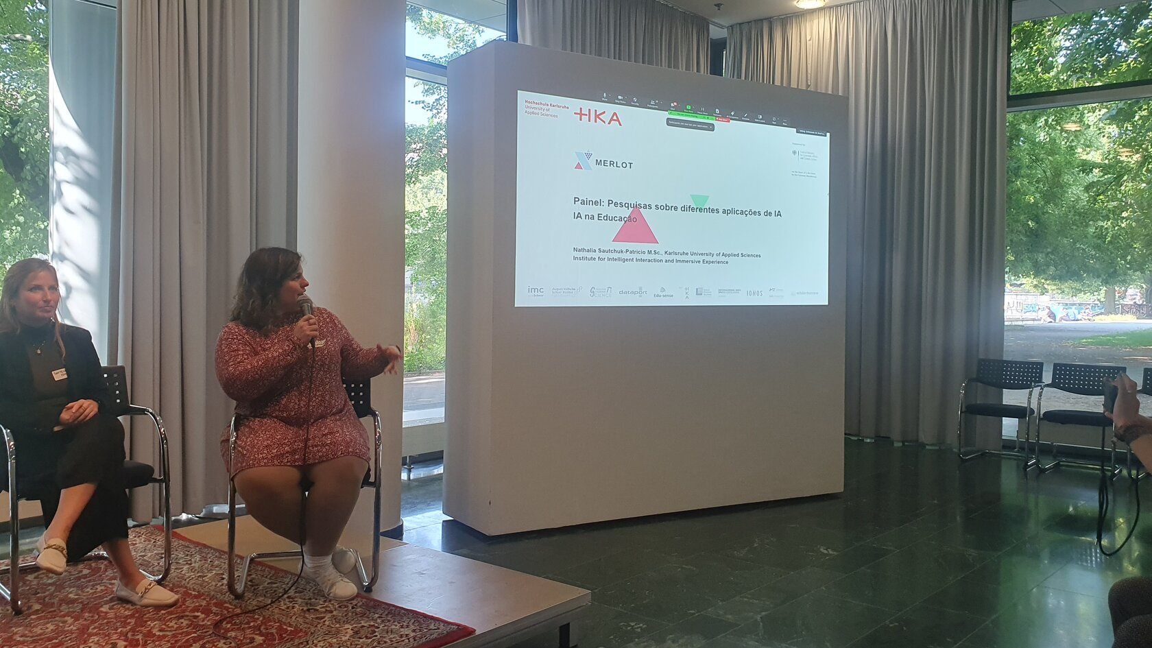 IIIX-Mitarbeiterin Nathalia Sautchuk Patricio sitzt neben einer Teilnehmerin auf dem Podium und stellt MERLOT auf einer Leinwand vor.