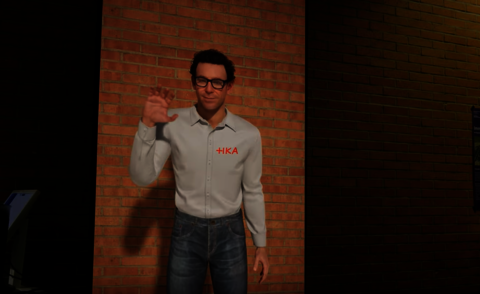 Auf dem Bild ist der Embodied Conversational Agent des IIIX zu sehen, dargestellt als lebensgro&szlig;er virtueller Charakter mit kurzen braunen Haaren, Brille, wei&szlig;em Hemd mit HKA-Logo und blauer Jeans. Der Agent steht vor einer Backsteinwand und hebt freundlich die Hand zum Gru&szlig;.