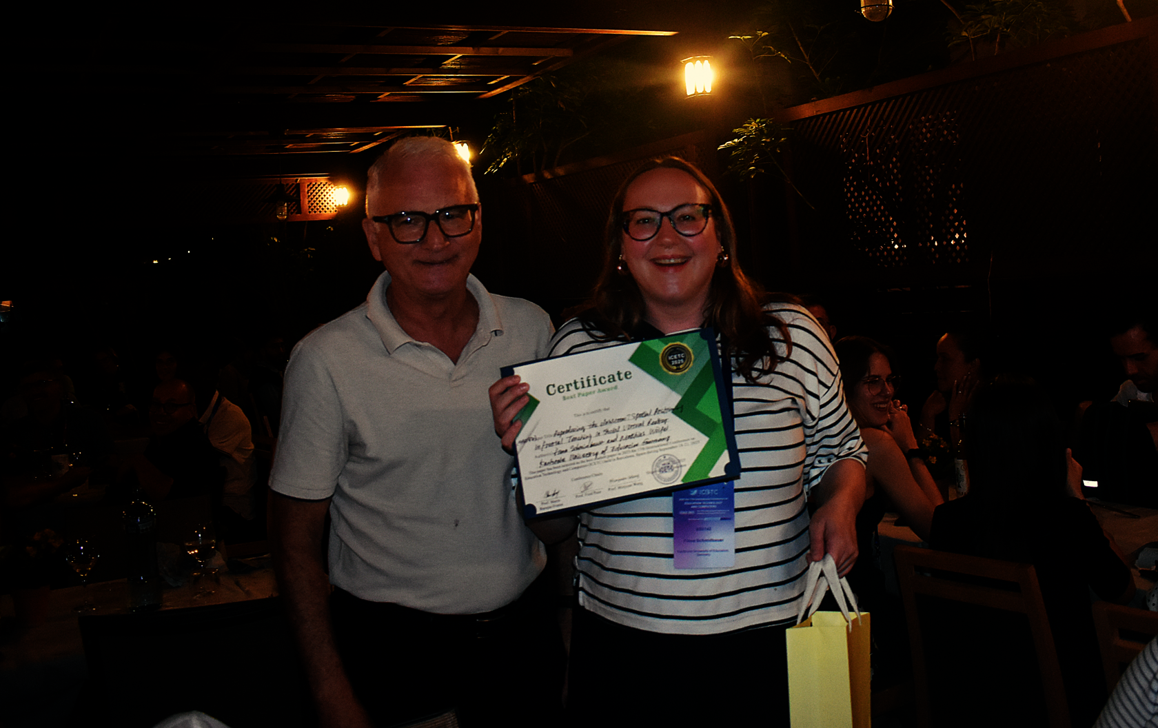 Promotionsstudentin Fiona Schmidbauer (IIIX) steht rechts neben dem Konferenzvorsitzenden Prof. Mario Barajas Frutos bei der Preisverleihung des Best Student Paper Award auf der ICETC 2025 und hält das Zertifikat für den Award in der Hand.