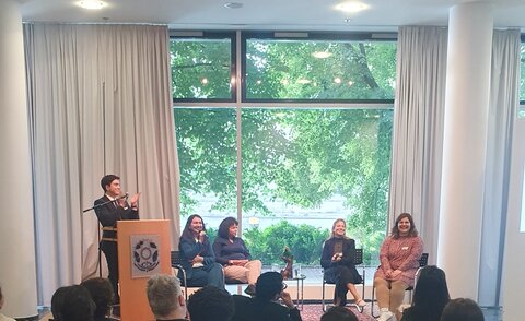 Vier Teilnehmerinnen sitzen in einer Diskussionsrunde, ein Teilnehmer steht hinter einem Redepult und klatscht.