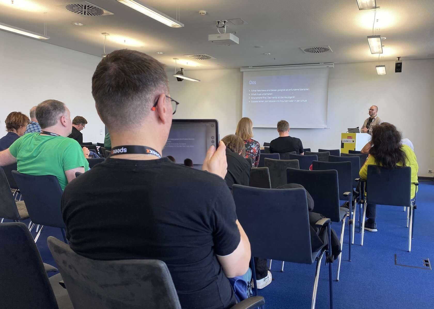 Participants at LEARNTEC 2026 follow a presentation in the congress program. In the foreground, a visitor is sitting with a tablet on which the content of the presentation is displayed. In the background, other members of the audience and Niklas Oesterle can be seen in front of a projection screen.