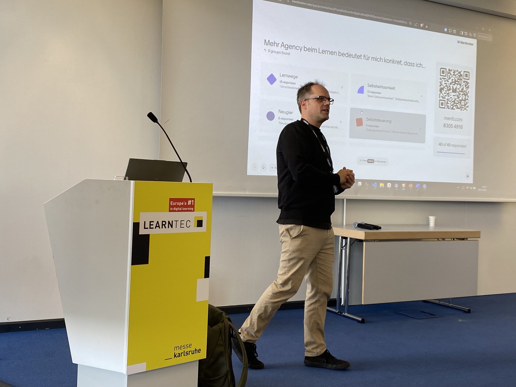 Thorsten Zylowski is standing next to a lectern labeled LEARNTEC 2026. Behind him is a screen on which his presentation slides can be seen. He looks attentively at the crowd.