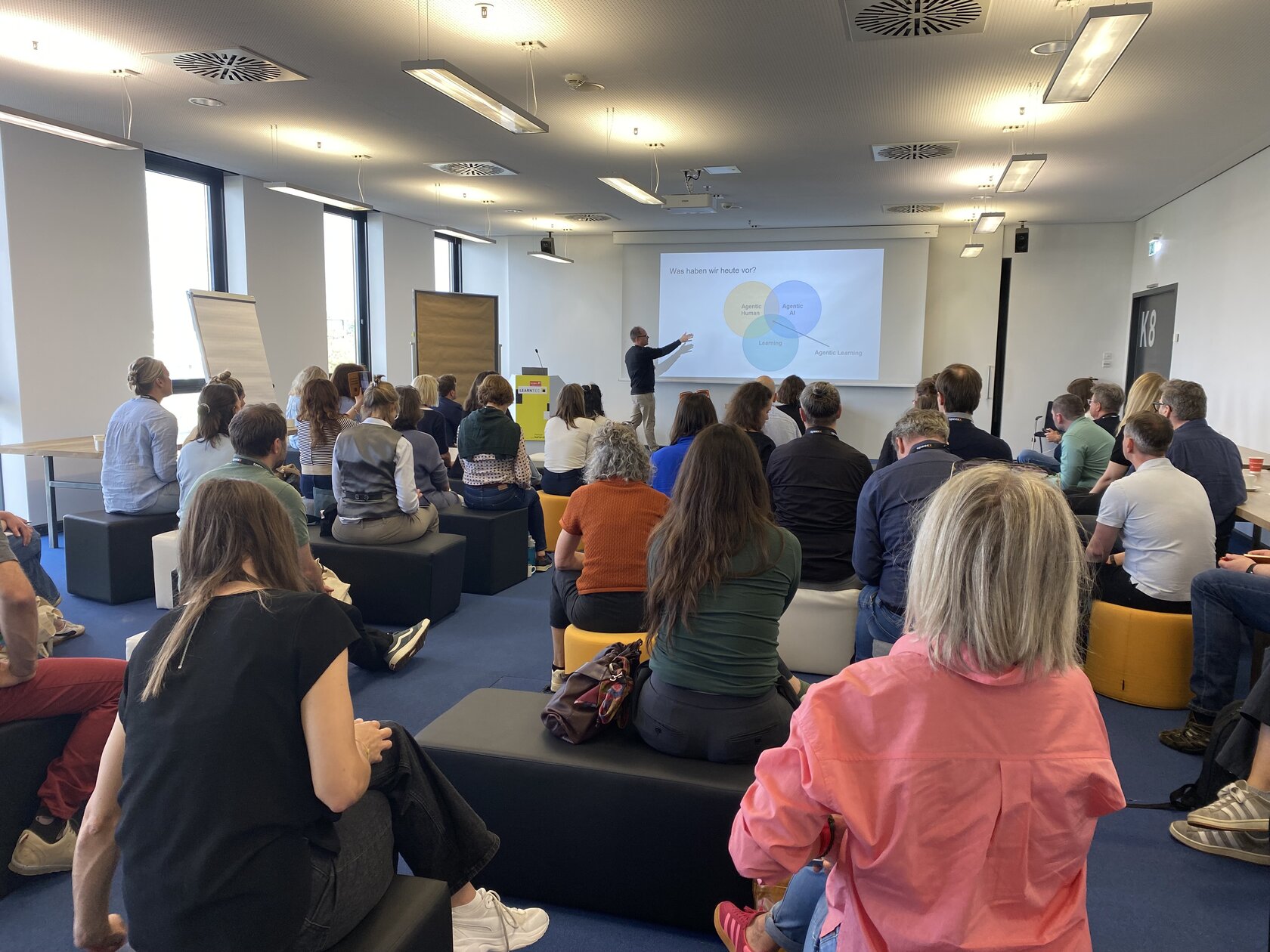 IIIX-Mitarbeiter Thorsten Zylowski steht vor einer großen Gruppe von Menschen, die auf Stühlen sitzen. Er zeigt mit seiner Hand auf eine Leinwand mit seinen Workshopfolien.
