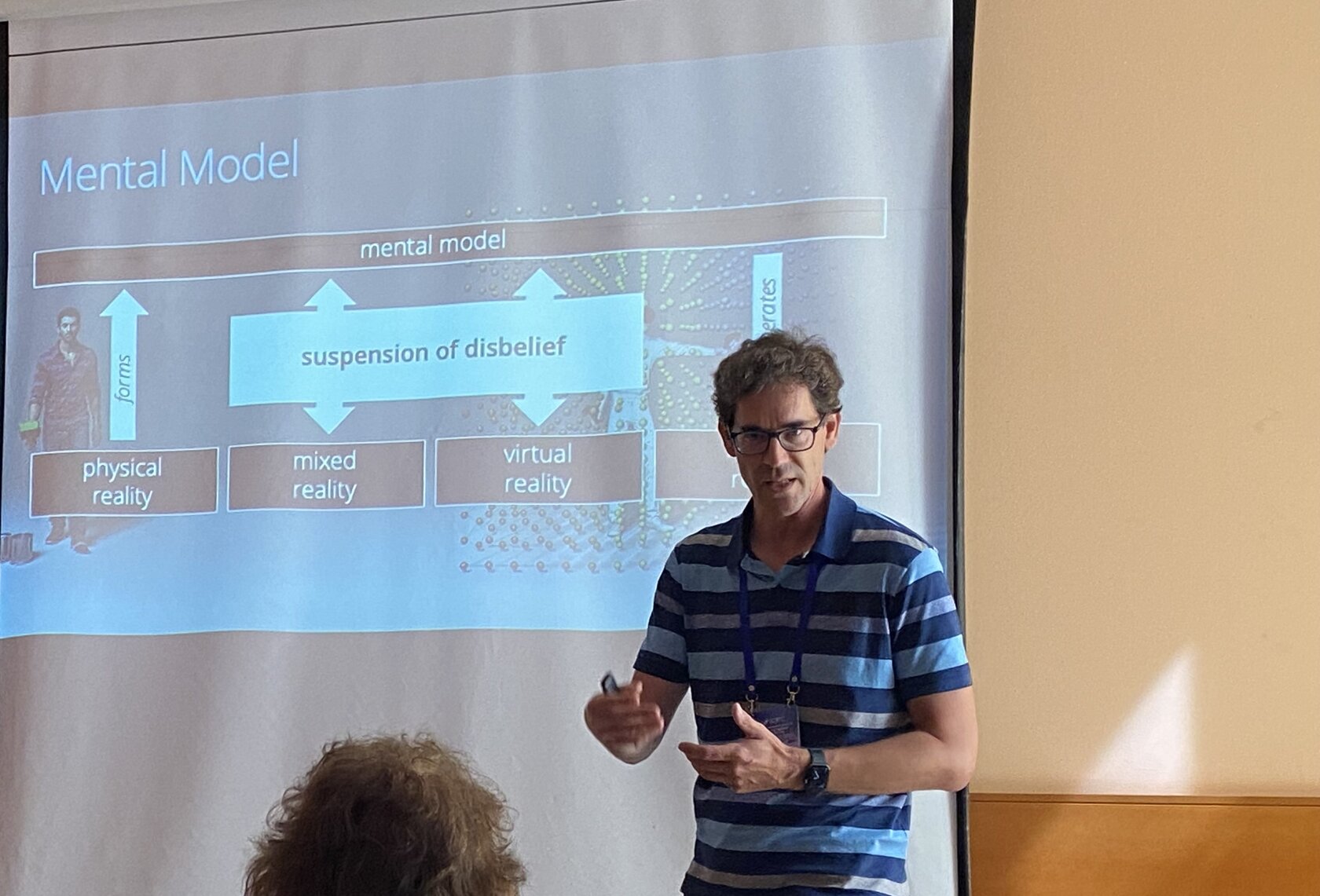 Prof. Dr. Matthias Wölfel steht in einem Vortragsraum vor einer Leinwand und spricht ins Mikrofon. Im Hintergrund ist eine Präsentationsfolie aus seiner Invited Speech zum Thema Virtual Reality und dem VR Campus zu sehen.